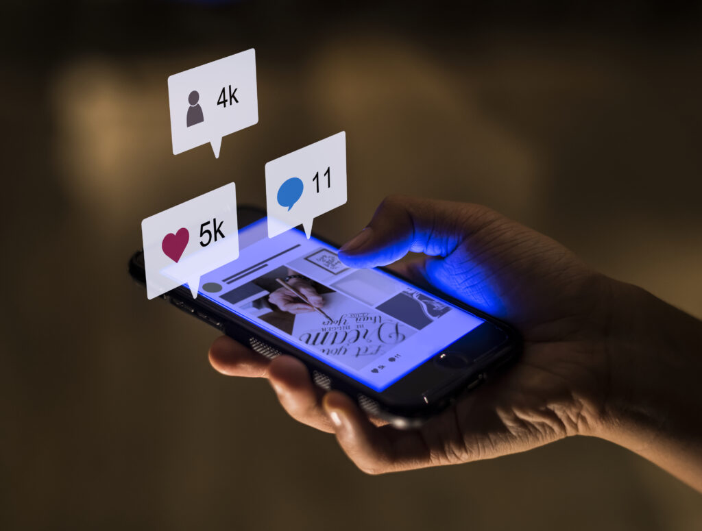 Uma pessoa olhando uma rede social. Há alguns ícones de curtidas, comentários e seguidores logo acima do celular