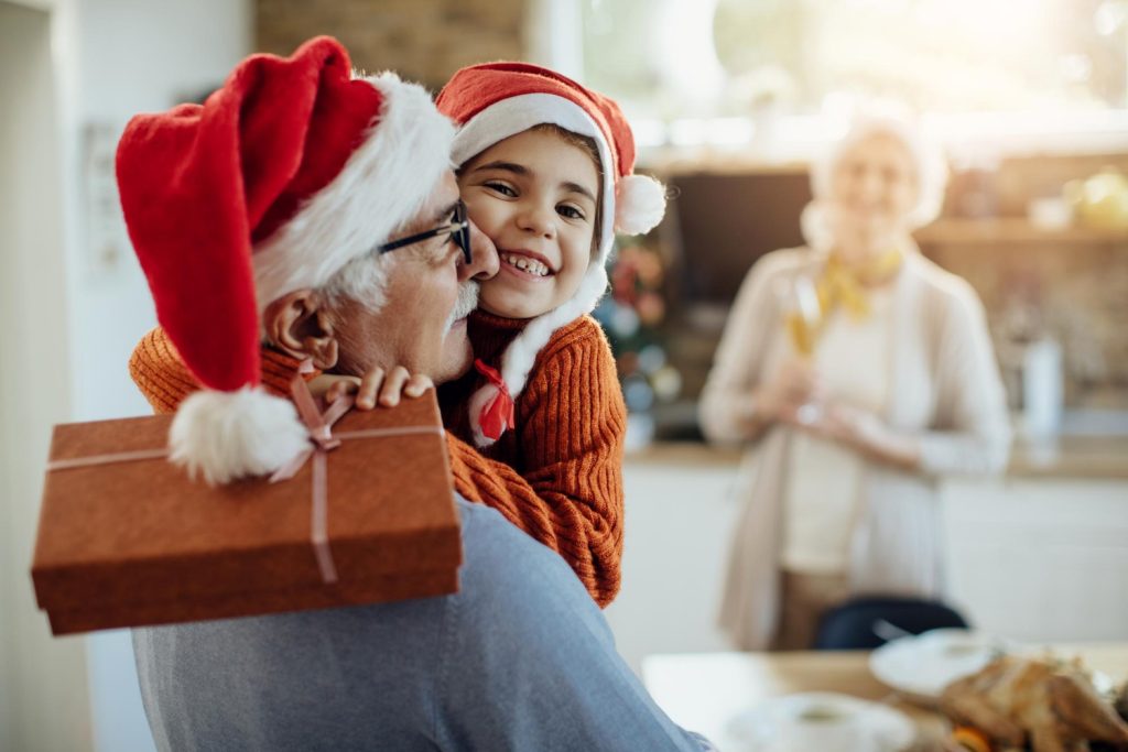 Neta abraçando o avô, em agradecimento pelo presente de natal que ganhou. A avó aparece no fundo, com um semblante feliz.
