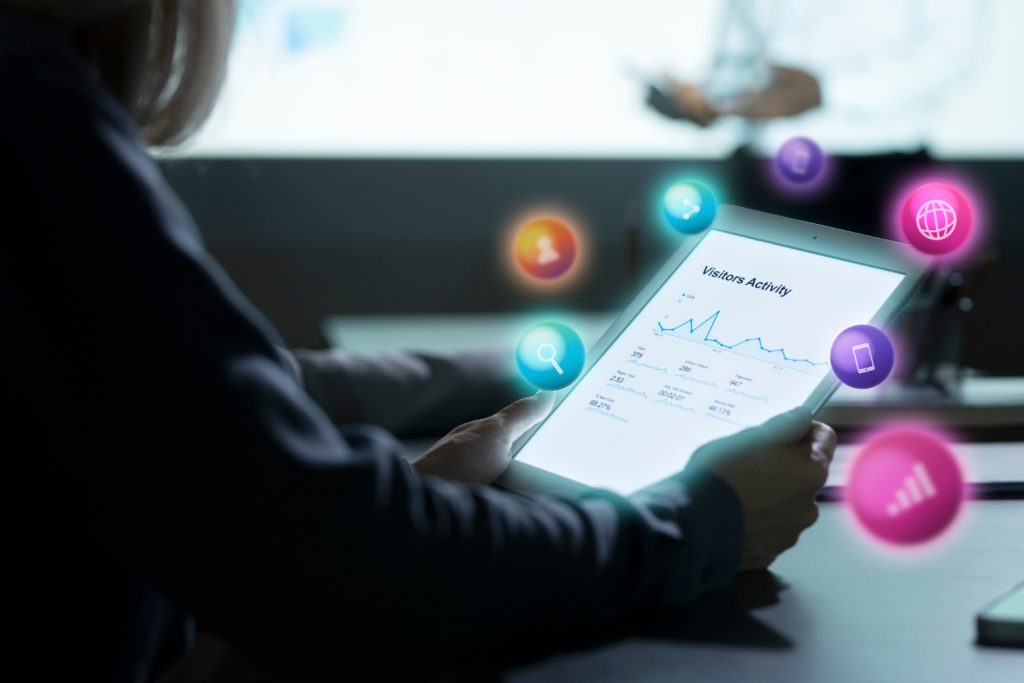 Pessoa olhando os resultados de uma estratégia digital no ipad. Tem alguns gráficos na tela, mostrando as atividades de visitantes no site. Há alguns ícones de ações do meio digital como pesquisa, compartilhamento, sinal de celular, meio que flutuando acima do ipad. 