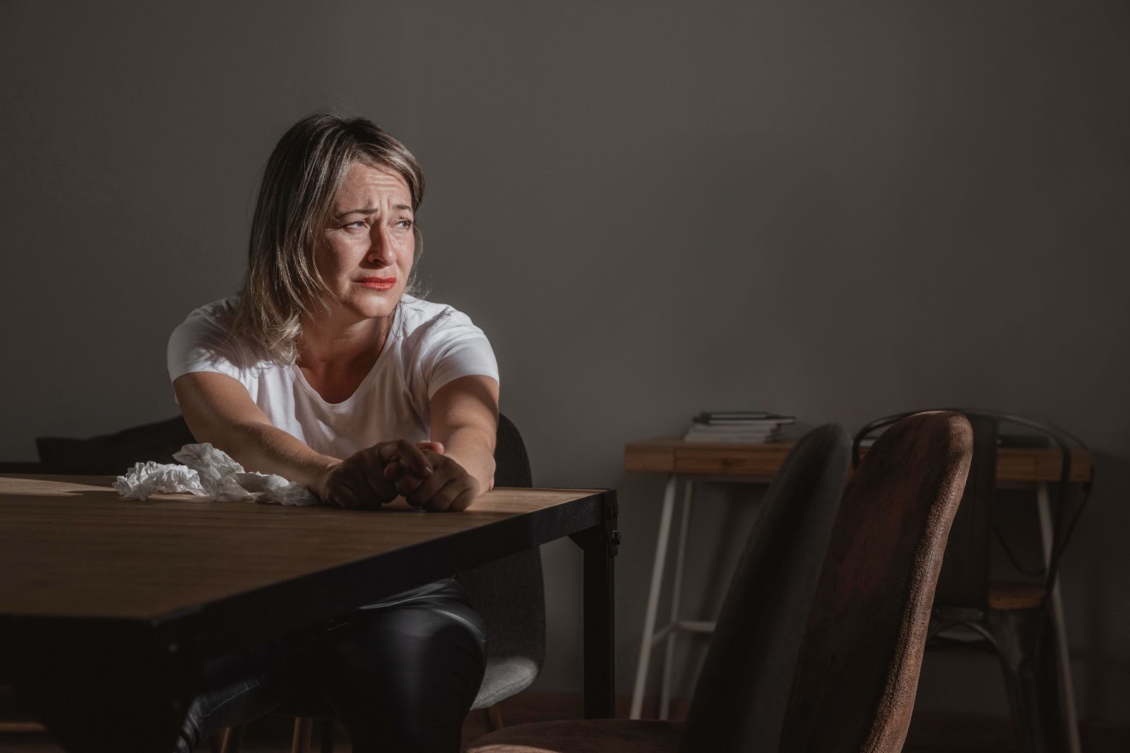 Mulher sentada à mesa, com as mãos unidas, lágrimas escorrendo pelo rosto, expressando tristeza profunda.